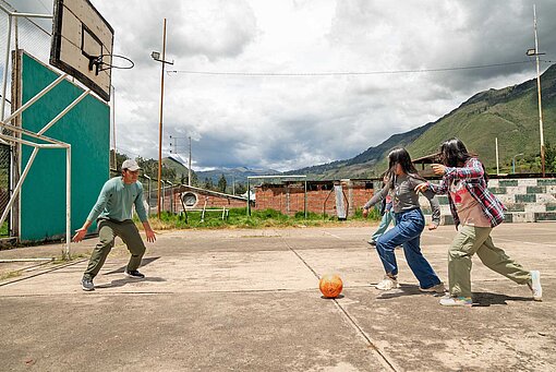 Ein sandiger Sportplatz, auf dem drei Mädchen gegen einen Mann Fußball spielen