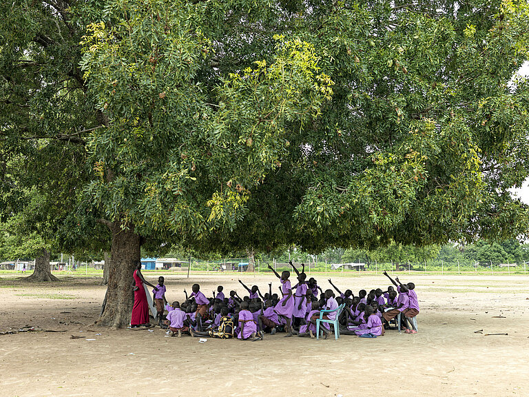 Eine Schulklasse steht mit einer Lehrerin unter dem Baum. Viele Kinder haben ihren Arm gehoben und melden sich.