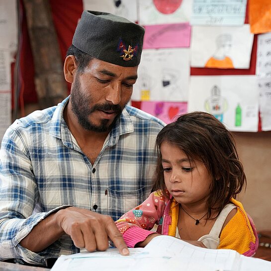 Vater sitzt neben seiner jüngsten Tochter und unterstützt sie beim Lesen eines Schulbuchs