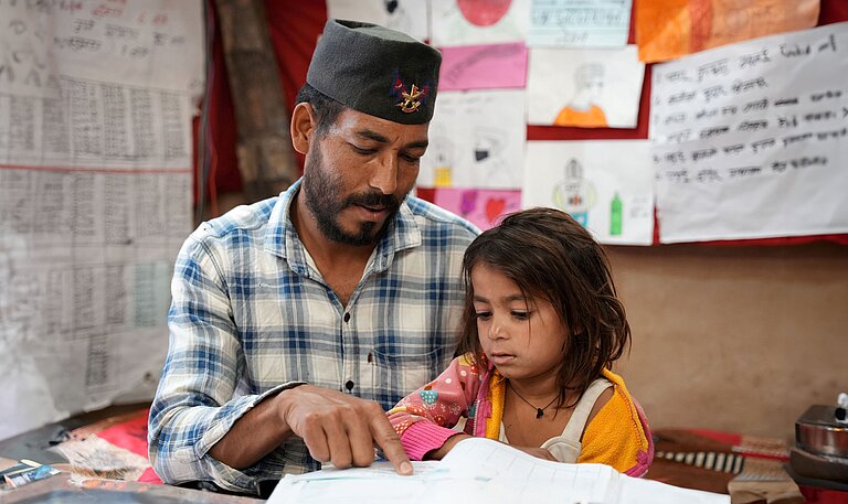 Vater sitzt neben seiner jüngsten Tochter und unterstützt sie beim Lesen eines Schulbuchs
