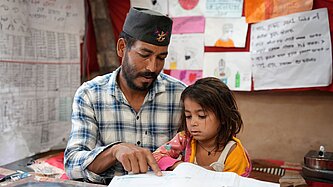 Vater sitzt neben seiner jüngsten Tochter und unterstützt sie beim Lesen eines Schulbuchs