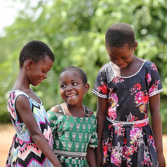 Drei lächelnde Kinder stehen gemeinsam im Freien und schauen sich an.