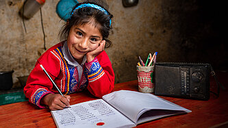 Lächelndes Mädchen in traditioneller Kleidung aus Peru macht Hausaufgaben an einem Holztisch mit Schreibheft, Bleistift und Radio.