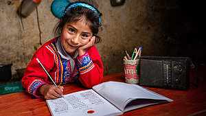 Lächelndes Mädchen in traditioneller Kleidung aus Peru macht Hausaufgaben an einem Holztisch mit Schreibheft, Bleistift und Radio.