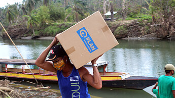 Eine Frau schleppt eine große Kiste mit Hygienekits auf dem Rücken. Im Hintergrund sieht man einen Fluss, auf dem ein leeres Kanu liegt