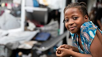 Mädchen lehnt an einem Balkongeländer und blickt leicht lächelnd in die Kamera