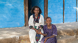 Eine Mutter und ihre Tochter in traditioneller Kleidung sitzen vor einem blauen Gebäude in Äthiopien und halten sich an den Händen. Sie sind ein Beispiel für die Zielgruppe des Projekts "Mädchen vor Gewalt schützen".