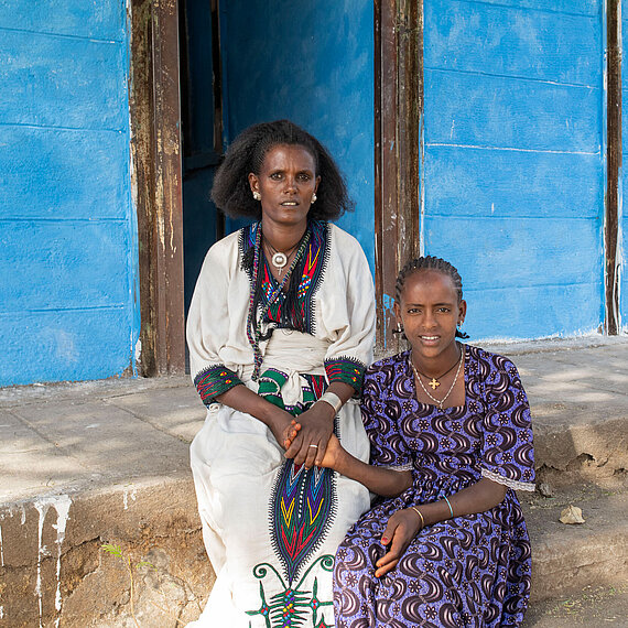 Eine Mutter und ihre Tochter in traditioneller Kleidung sitzen vor einem blauen Gebäude in Äthiopien und halten sich an den Händen. Sie sind ein Beispiel für die Zielgruppe des Projekts "Mädchen vor Gewalt schützen".