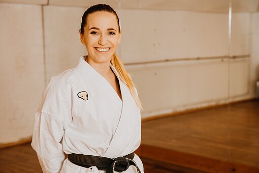 Eine Frau trägt einen Karate-Anzug mit schwarzem Gürtel und schaut in die Kamera