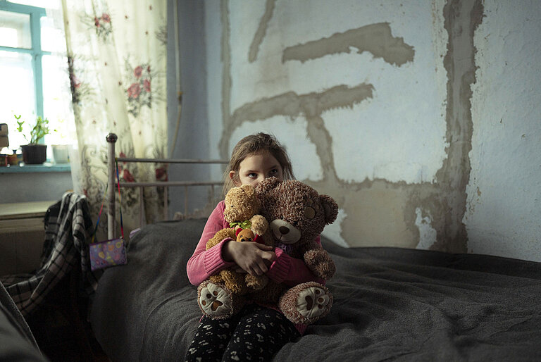 Ein junges Mädchen sitzt auf einem Bett und hält zwei Teddys im Arm, an die sie ihr Gesicht schmiegt.