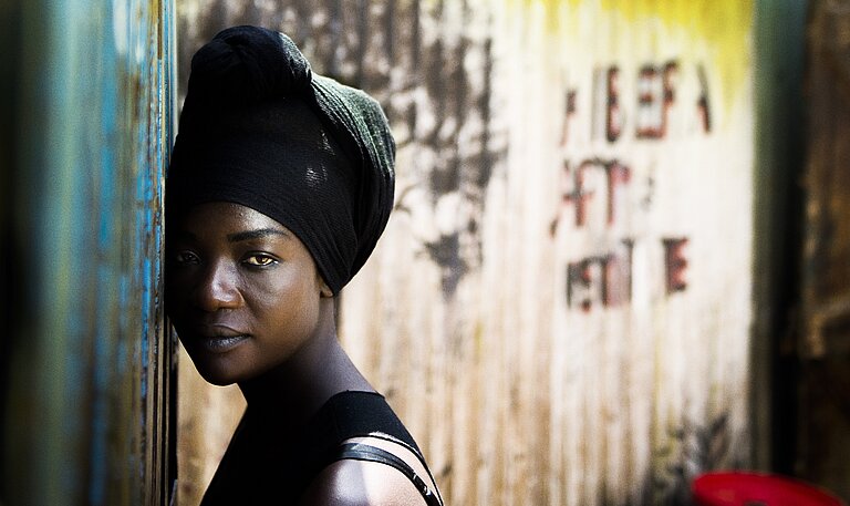 Eine afrikanische Frau, die ihre Haare in ein Tuch gebunden ha, lehnt mit dem Gesicht an einer Wellblechwand und schaut in die Kamera.
