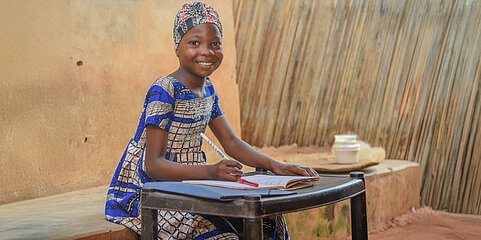 Ein Mädchen mit traditioneller, bunter Kleidung sitzt an einem kleinen Tisch. Vor ihr liegt ein Heft und sie hält einen Stift in der Hand. Sie lächelt in die Kamera.
