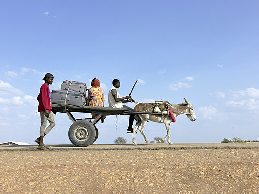 Zwei sudanesische Menschen sitzen auf einem Karren, der von einem Esel gezogen wird, ein weiterer Mensch läuft nebenher