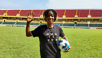 Eine junge Schwarze Frau steht mitten in einem Fußballstadion. Sie winkt in die Kamera und hat einen Ball im Arm.