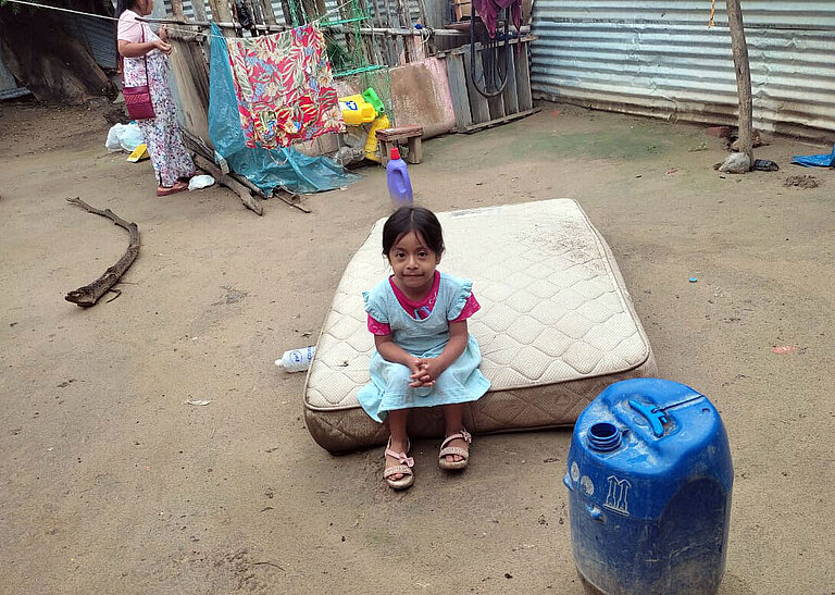Ein kleines Mädchen sitzt im Hof auf einer Matratze, die auf dem schlammigen Boden liegt.