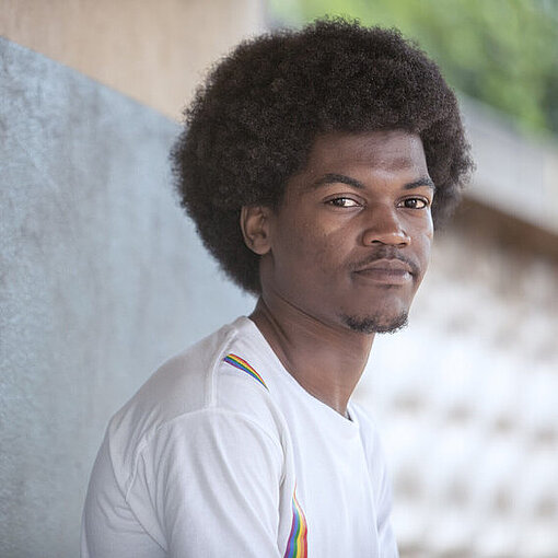 Portraitfoto von Custodio. Er trägt ein weißes Shirt und lehnt gegen eine graue Wand. 