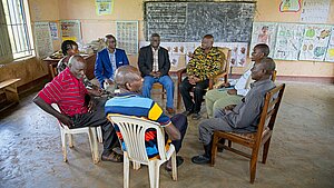 Vätergruppe im Baba Club bei einem Treffen im ECD-Zentrum im Distrikt Kamuli, Uganda. Die Männer sitzen im Stuhlkreis in einem Klassenzimmer, um über Bildungsmöglichkeiten und Ernährung für Schulkinder zu sprechen.