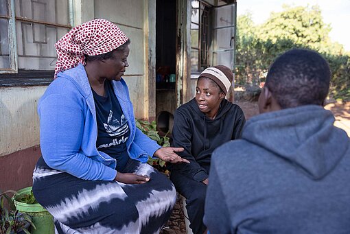 Zwei Frauen und ein Mann sitzen auf Hockern vor einem grün gestrichenen Haus