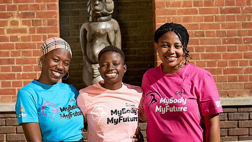 Drei junge Frauen aus Simbabwe halten sich in den Armen und lachen. Sie tragen T-Shirts mit der englischen Aufschrift "Mein Körper, meine Zukunft".