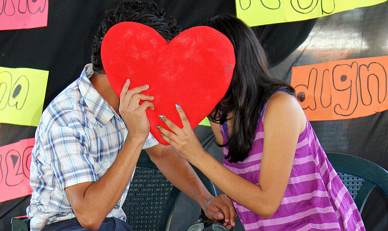 Ein Junge und ein Mädchen sitzen auf Stühlen und stecken die Köpfe zusammen, vor ihre Gesichter halten sie ein Herz aus Pappe.