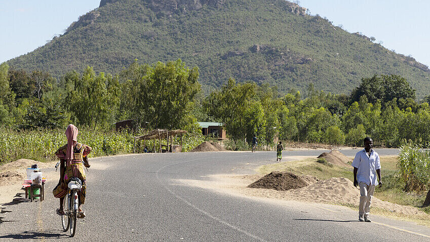 Asphaltierte Straße im Bezirk Phalombe, Malawi Eine asphaltierte Straße mit bewaldetem Hügel im Hintergrund. Auf der Straße fährt eine Frau Fahrrad und ein Mann geht spazieren.