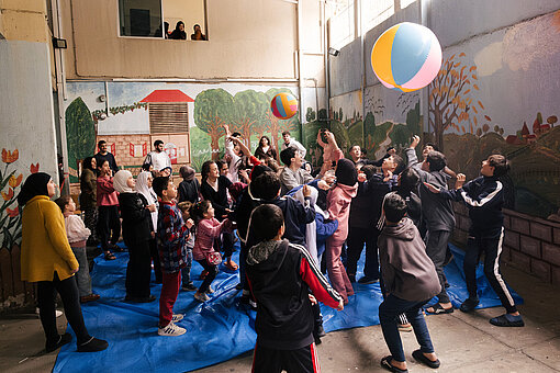 Kinder und Erwachsene spielen in einem kinderfreundlichen Raum mit bunten Bällen. Die Wände sind mit Malereien dekoriert, auf denen Häuser und Natur zu sehen sind.