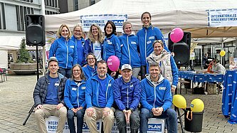 Mehrere Frauen und Männer in hellblauebn Jacken mit dem Plan-Logo sitzen vor einem weißen Festzelt