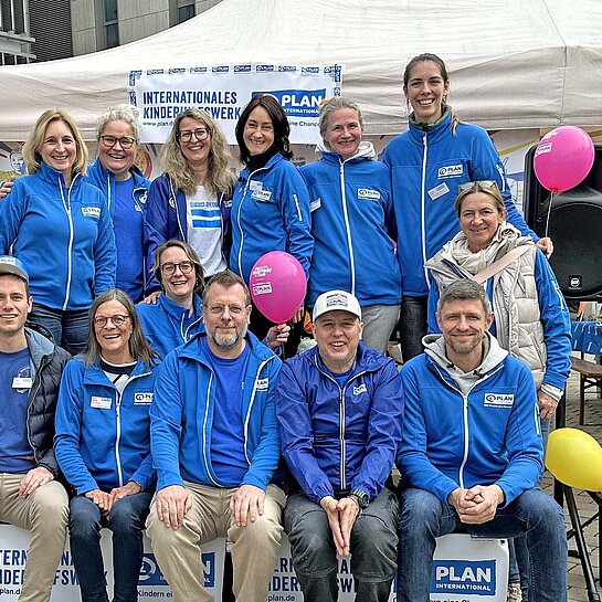 Mehrere Frauen und Männer in hellblauebn Jacken mit dem Plan-Logo sitzen vor einem weißen Festzelt