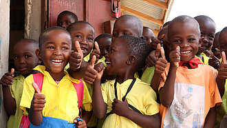 Gruppe lachender Schulkinder in gelben Uniformen zeigt Daumen hoch.