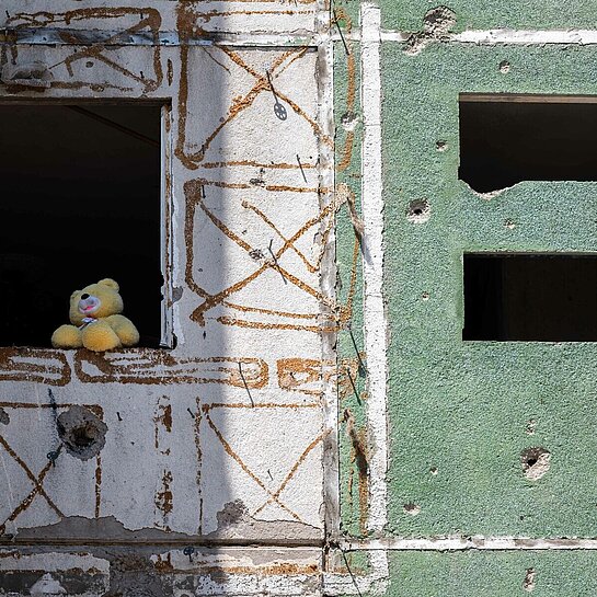 Eine Häuserfront mit Einschusslöchern, in einem Fenster sitzt ein Plüsch-Teddy