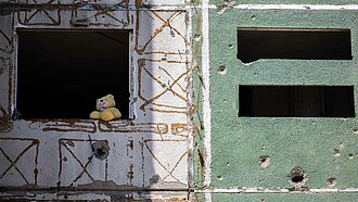 Eine Häuserfront mit Einschusslöchern, in einem Fenster sitzt ein Plüsch-Teddy