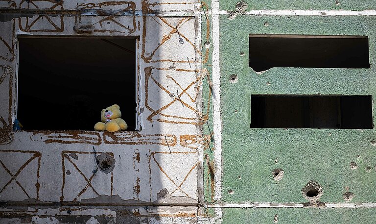 Eine Häuserfront mit Einschusslöchern, in einem Fenster sitzt ein Plüsch-Teddy