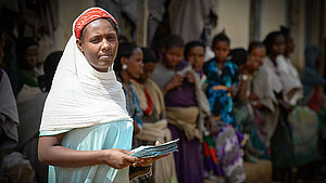 Eine Frau hält ein Stapel Dokumente in der Hand und blickt in die Kamera. Im Hintergrund stehen mehrere weitere Frauen. Sie ist ein Beispiel für die Zielgruppe des Projekts "Mädchen vor Gewalt schützen".