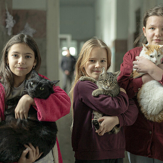 Drei Mädchen stehen in einem Flur und lächeln in die Kamera. Jede von ihnen hat eine Katze auf dem Arm