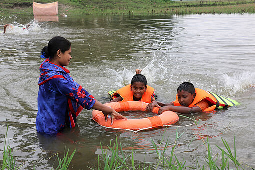 Irin steht im Teich. Neben ihr schwimmen zwei Jungen in Schwimmwesten, die sich an einem Rettungsring festhalten.