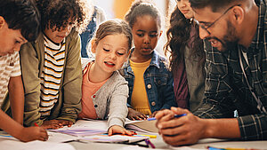  Mehrere Kinder arbeiten gemeinsam mit einem Lehrer an einem Tisch voller Malstifte und Papier