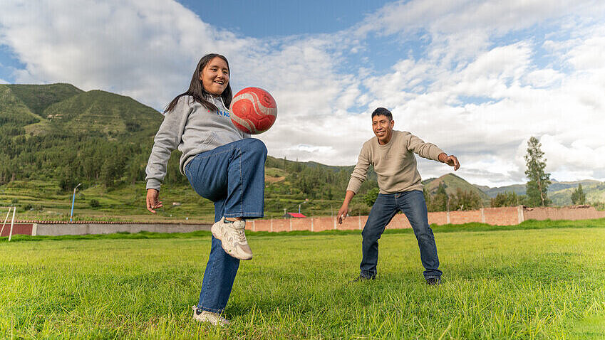 Mitzy spielt Fußball auf einer Wiese Ein Mädchen und ein Junge spielen zusammen Fußball auf einer Wiese. Hinter ihnen ragen Berge empor.