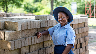 Junges Mädchen in Schuluniform steht lächelnd vor einem Stapel Ziegelsteine