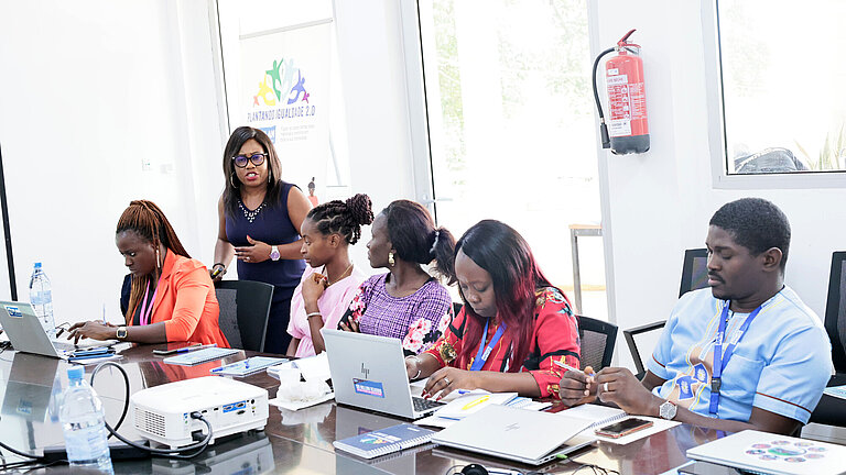 An einem großen Tisch sitzen fünf Menschen an Laptops, es steht ein Beamer auf dem Tisch und eine Frau trägt den Workshop-Teilnehmenden etwas vor