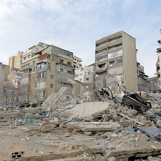 Eine zerstörte Wohngegend in der Stadt mit eingestürzten Gebäuden und Trümmern. Mehrere halb intakte Hochhäuser stehen im Hintergrund.