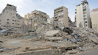 Eine zerstörte Wohngegend in der Stadt mit eingestürzten Gebäuden und Trümmern. Mehrere halb intakte Hochhäuser stehen im Hintergrund.