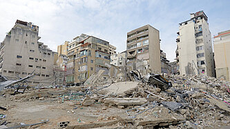 Eine zerstörte Wohngegend in der Stadt mit eingestürzten Gebäuden und Trümmern. Mehrere halb intakte Hochhäuser stehen im Hintergrund.
