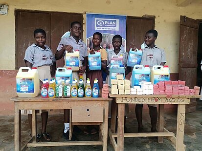 Fünf ghanaische Schulkinder präsentieren stolz selbst hergestellte Hygieneprodukte wie Flüssigseife, Waschmittel und feste Seifenstücke. Die Produkte sind auf Tischen ausgestellt, im Hintergrund ist ein Banner von Plan International zu sehen mit dem Slogan „Until we are all equal“.