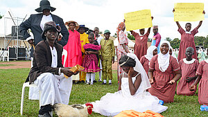 Schulaufführung in Uganda: Kinder stellen Kinderehe dar, Mädchen im Brautkleid sitzt traurig auf dem Boden, andere Kinder halten Protestplakate gegen Vergewaltigung und fehlende Hygieneprodukte.
