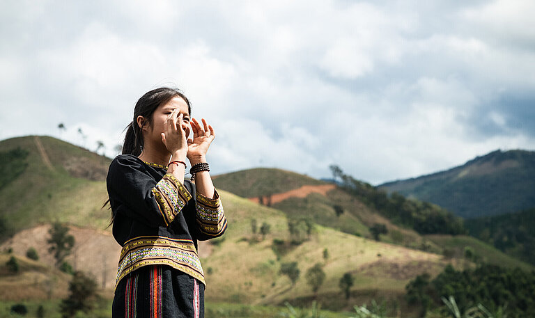 Ein Mädchen aus Vietnam steht draußen und hält beide Hände vor den Mund, als würde es etwas rufen