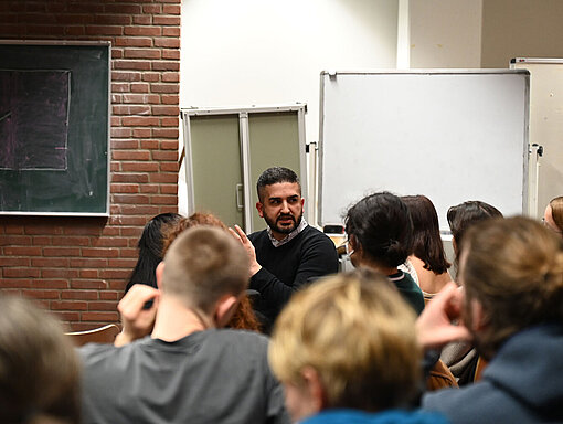 Ein Mann sitzt im Publikum im Hörsaal und spricht mit den anderen. 