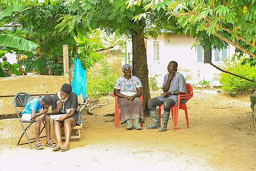 Wie viele Frauen in Ghana sah sich Paulina (2. v. r.) mit systembedingten Hindernissen konfrontiert Zwei Kinder und zwei Erwachsene sitzen unter einem Baum vor einem Haus