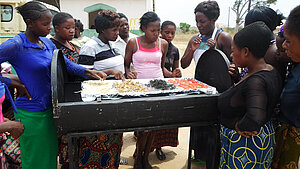 Junge Frauen in Sambia lernen den Einsatz eines Solartrockners zur nachhaltigen Lebensmittelverarbeitung. Eine Gruppe junger Frauen in Sambia steht um einen Solartrockner, um verschiedene Lebensmittel wie Tomaten, grünes Blattgemüse und Samen zu trocknen – Teil eines Bildungsprojekts zum Klimawandel und zur Ernährungssicherheit.