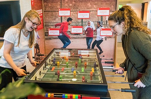 Dürfen Mädchen und Frauen überall auf der Welt Fußball spielen? Antworten liefert die interaktive Schau „Mission 2030“ Zwei junge Frauen stehen an einem Fußball-Kicker-Tisch, der Teil der Erlebnisausstellung von Plan International ist