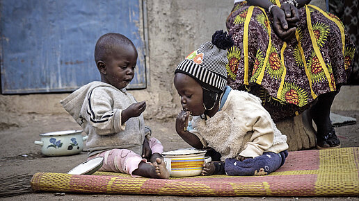Zwei Kleinkinder essen auf dem Boden auf einer Decke aus einer Schüssel Brei.